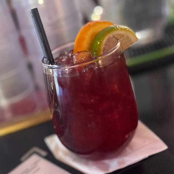 A red cocktail with ice, garnished with orange and lime slices, and a black straw.
