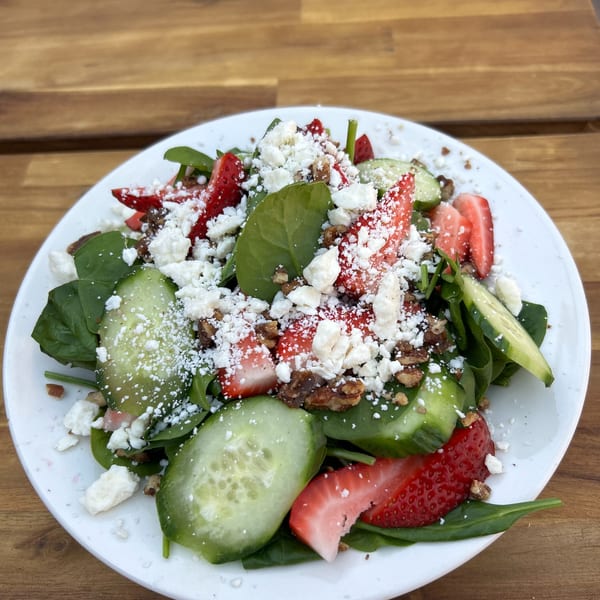 A fresh spinach salad with strawberries, cucumbers, feta cheese, and nuts on a white plate.